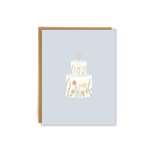 A white, two tiered cake with floral decorations, against an elegant blue backdrop